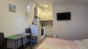 A compact room with a bed, a desk with a chair, a kitchenette, and a TV mounted on the wall. - Studio Apartment Nia (Nin)