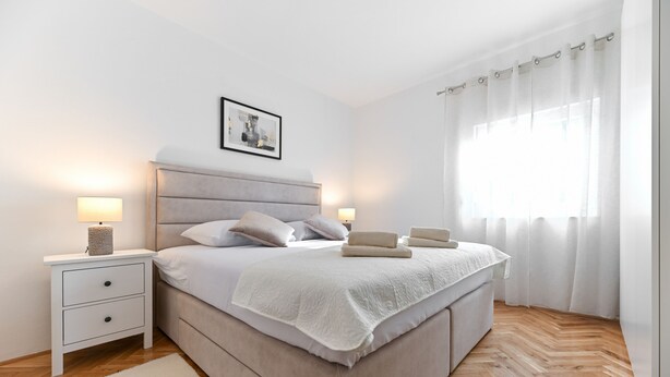 Une chambre à coucher moderne avec un grand lit, des lampes de chevet, un tableau encadré au mur et une fenêtre avec des rideaux légers.