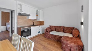 Een compacte woonruimte met kitchenette, een bruine gestreepte bank, een eettafel met stoelen en een witte muur met een lamp.