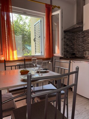 Un coin repas avec une table dressée pour deux personnes, des chaises, une fenêtre avec des rideaux et un espace cuisine équipé d’une cuisinière et de placards.