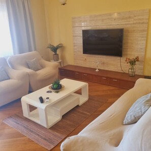 Ett modernt vardagsrum med en vit soffa, ett vitt soffbord, en platt-TV som är monterad på en väggpanel i trä och en liten krukväxt.