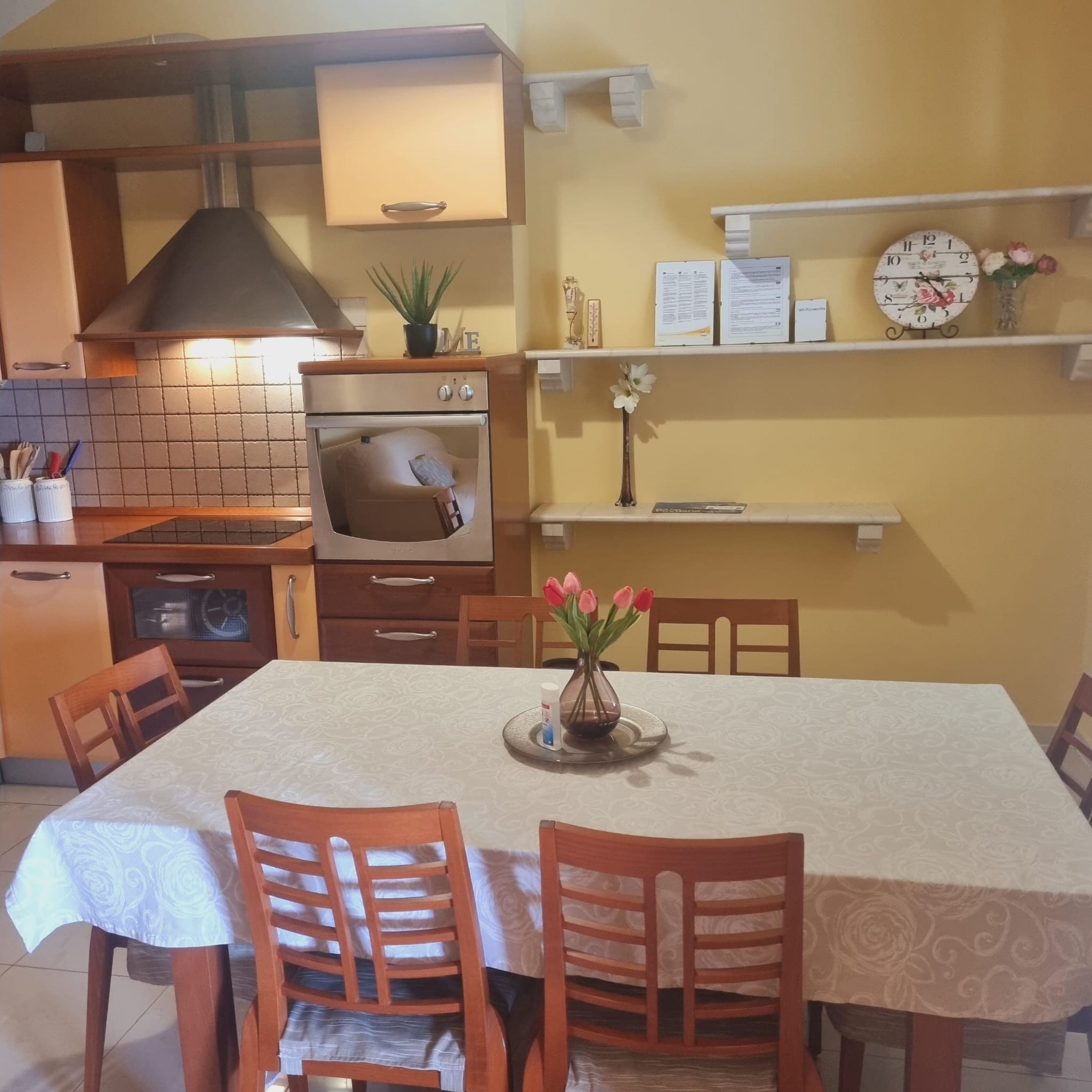 A kitchen with a stove, oven, and a dining table with chairs. There is a clock on the wall and a shelf with items.