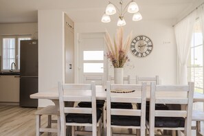 Ein Essbereich mit einem weißen Tisch und Stühlen, einer Vase mit getrockneten Pflanzen, einer Wanduhr und einer Küche im Hintergrund.