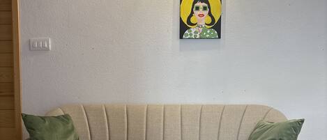 Un sofá beige con dos cojines verdes, una lámpara de pared y una obra de arte enmarcada encima.