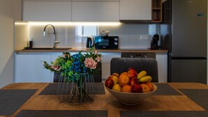 A modern kitchen with white cabinets, a microwave, and a refrigerator. A vase with flowers and a bowl of fruit are on the dining table. - Apartment Vista Mare 2 With Heated Pool (Sveti Filip i Jakov)