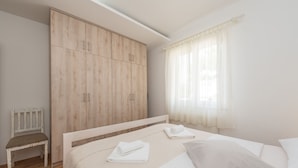 A bedroom with a bed, a wooden wardrobe, a chair, and a window with curtains. - Apartment Brevilacqua (Privlaka)