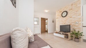 A living room with a brown sofa, a television on a wooden stand, a clock on the wall, and a potted plant. - Apartment Brevilacqua (Privlaka)