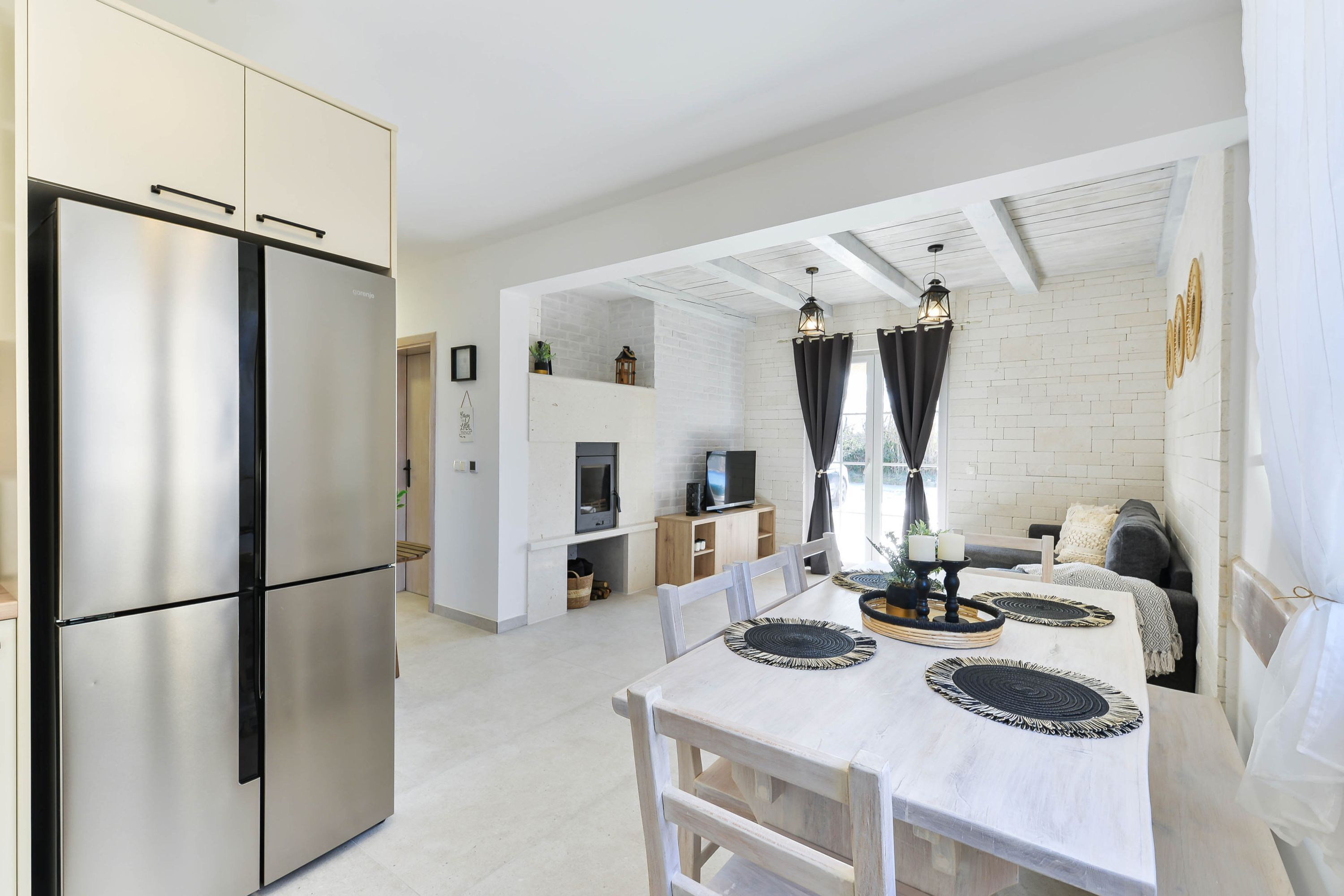 A modern kitchen with a stainless steel refrigerator, a dining table with black and white placemats, and a cozy seating area with a sofa and curtains.