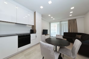 A modern kitchen with white cabinets and black appliances, a dining area with a dark table and white chairs, and a dark sofa in a living area with a large window. - Luxe Apartment Verto (Zadar)