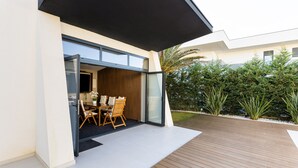 A modern house with a patio area featuring a wooden deck, a glass door leading to an interior space with a dining table and chairs, and a well-maintained garden with various plants. - Villa Positiva With Heated Pool (Vir)