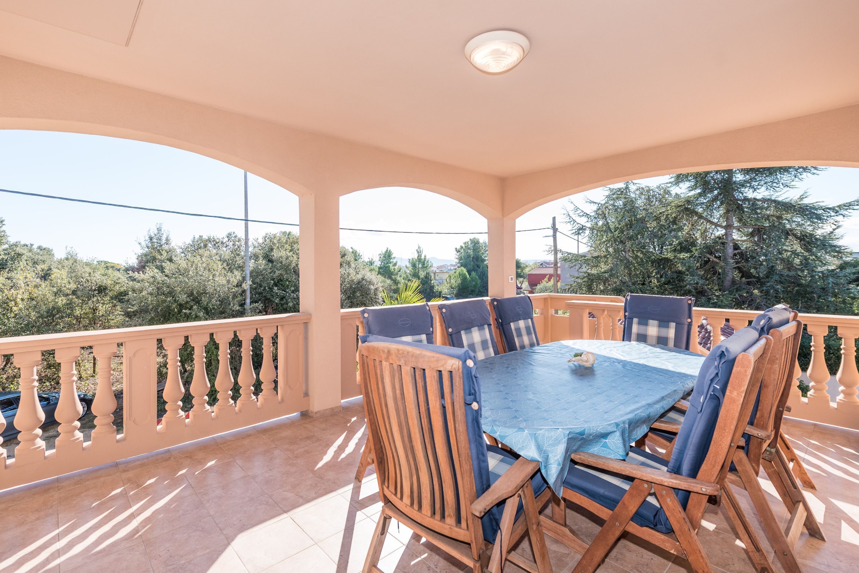 Un espacio de comedor al aire libre techado, con una mesa, sillas, barandilla y vistas a árboles y edificios.
