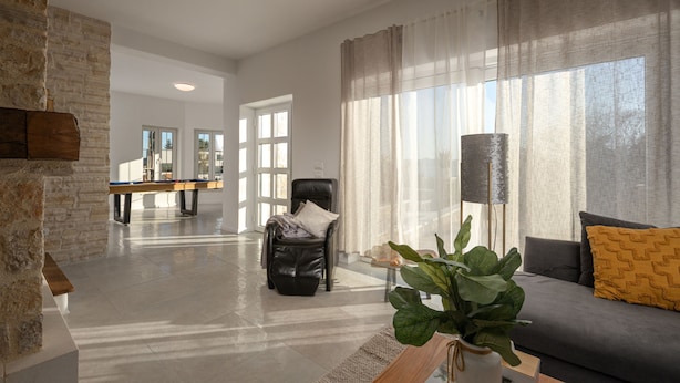 Ein geräumiges Wohnzimmer mit Billardtisch, Ledersessel, einer Couch mit Kissen und einem großen Fenster mit durchsichtigen Vorhängen.