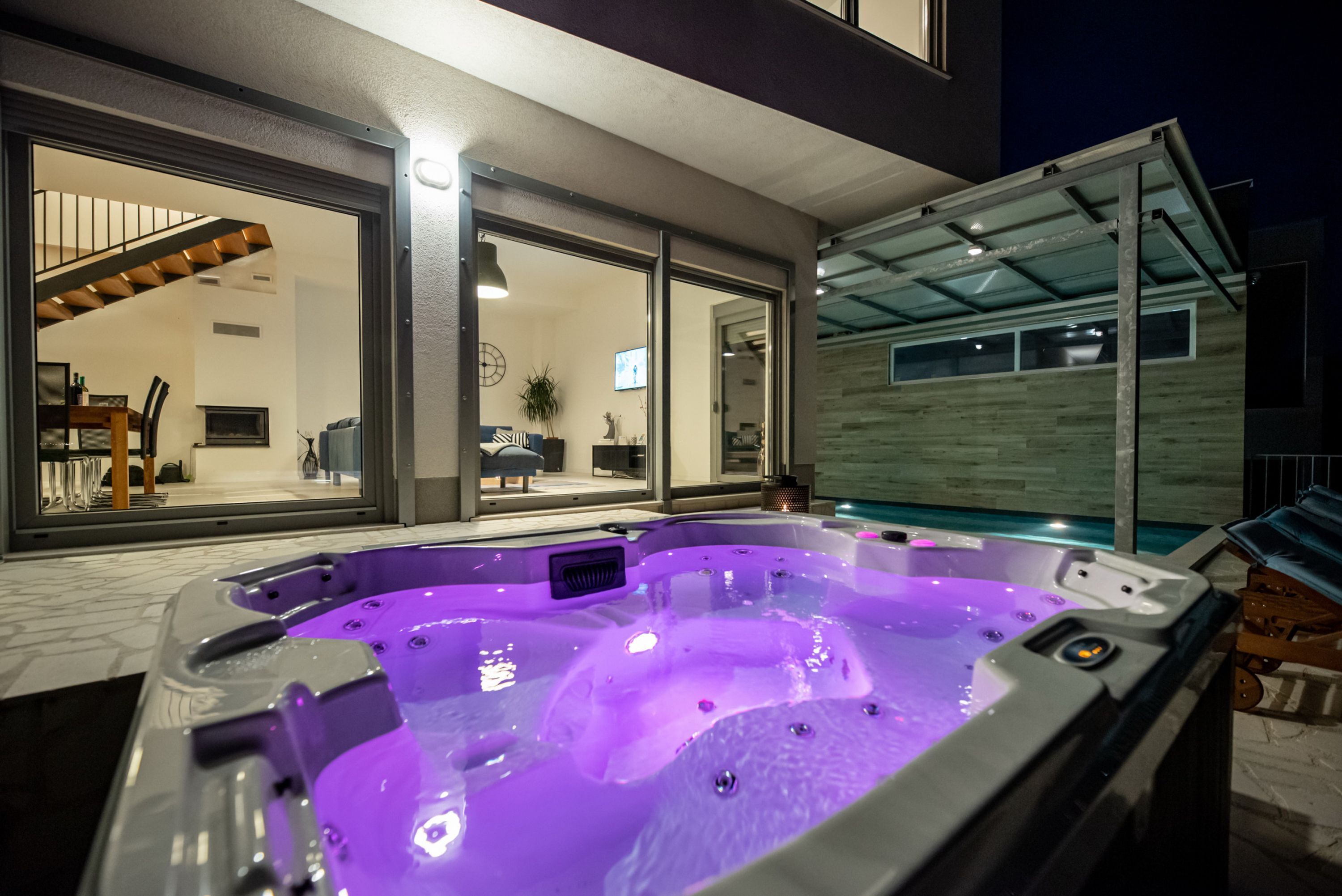 Ein moderner Außenbereich mit Whirlpool, Glasumrandung, Holzterrasse und Blick zum Wohnbereich.