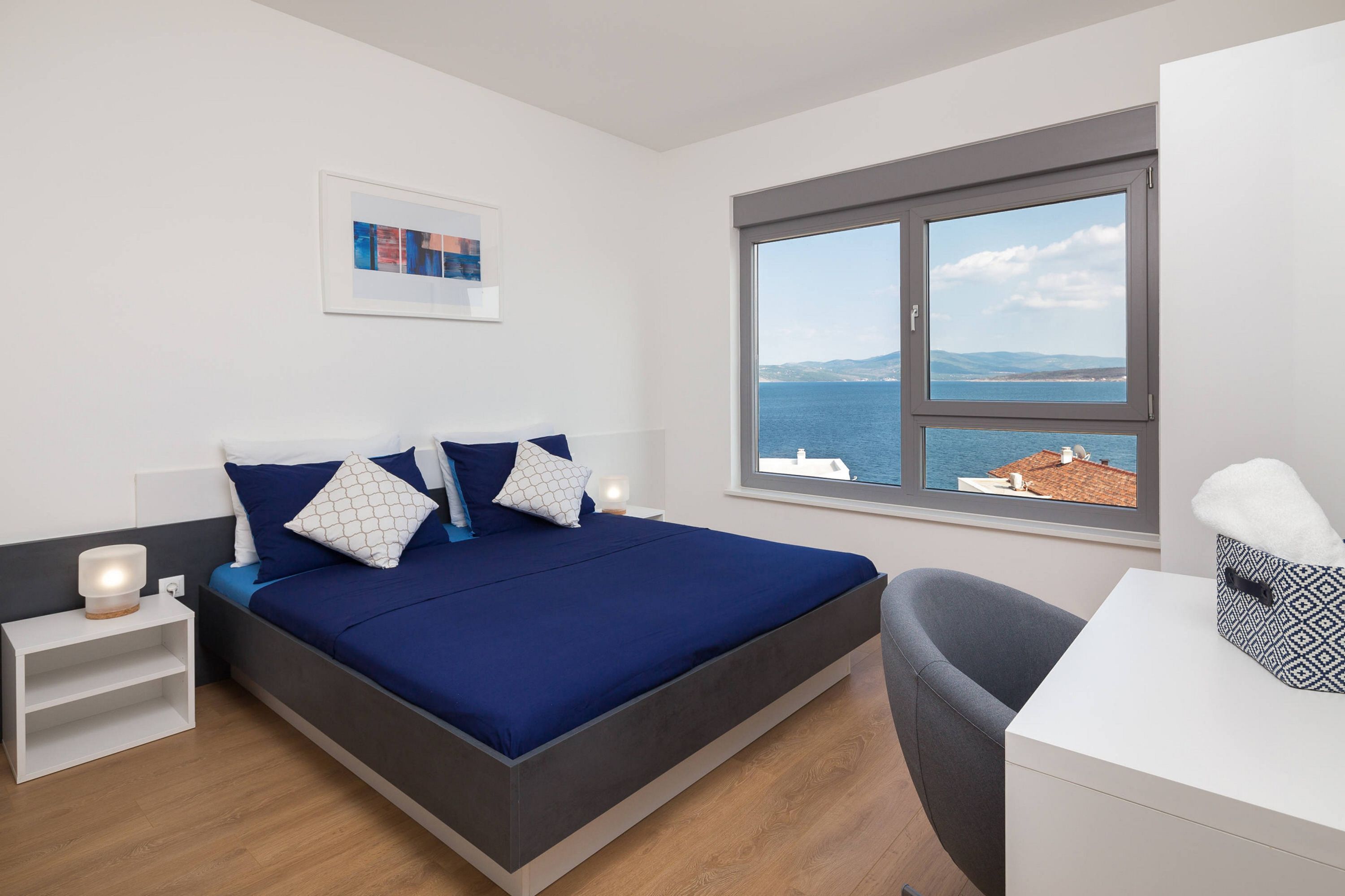 A modern bedroom with a large bed, a desk, and a window with a view of the sea.