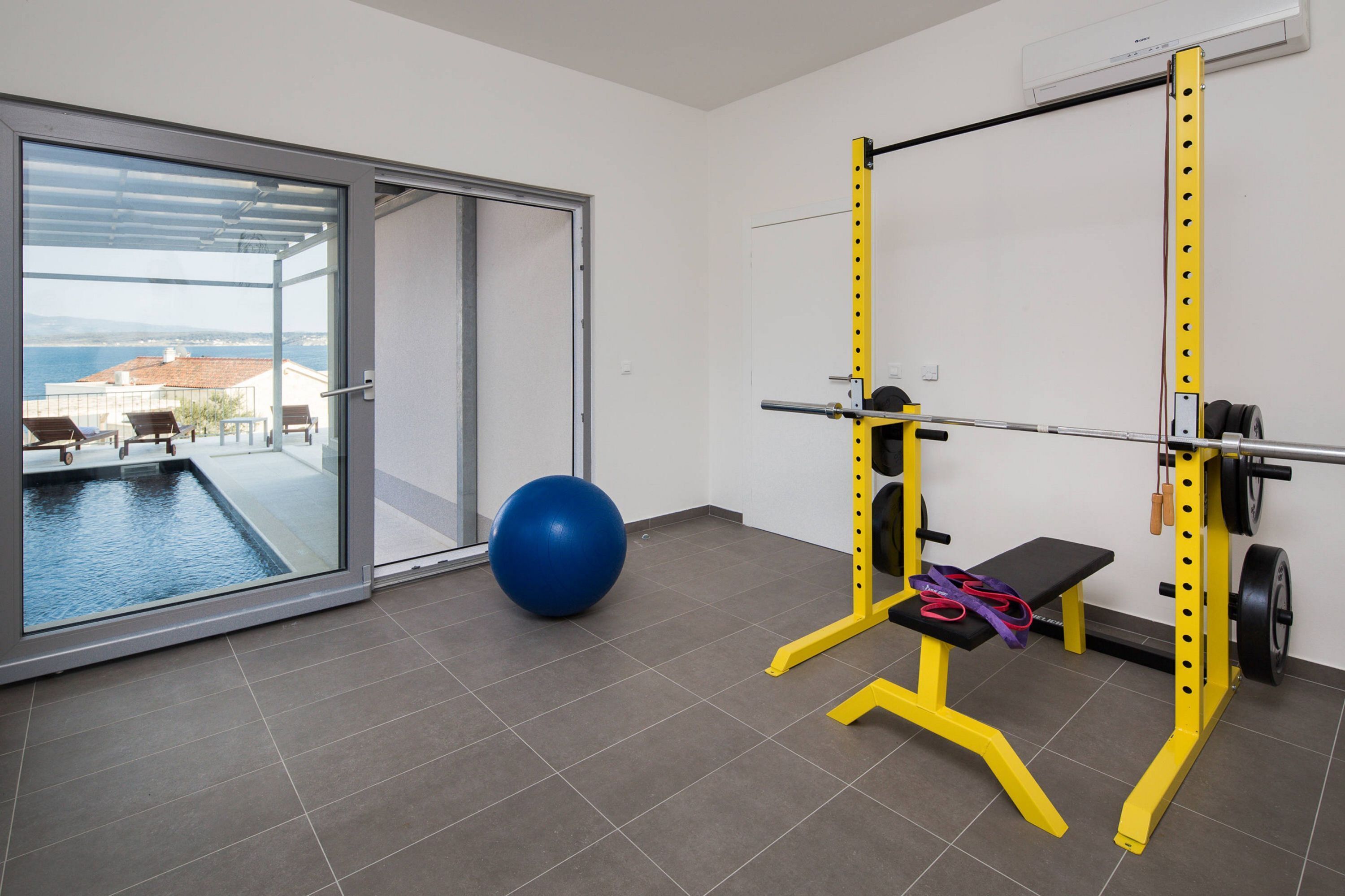 A gym with a squat rack, a blue exercise ball, and a bench. There is a sliding glass door leading to a balcony with a view of the sea and buildings.