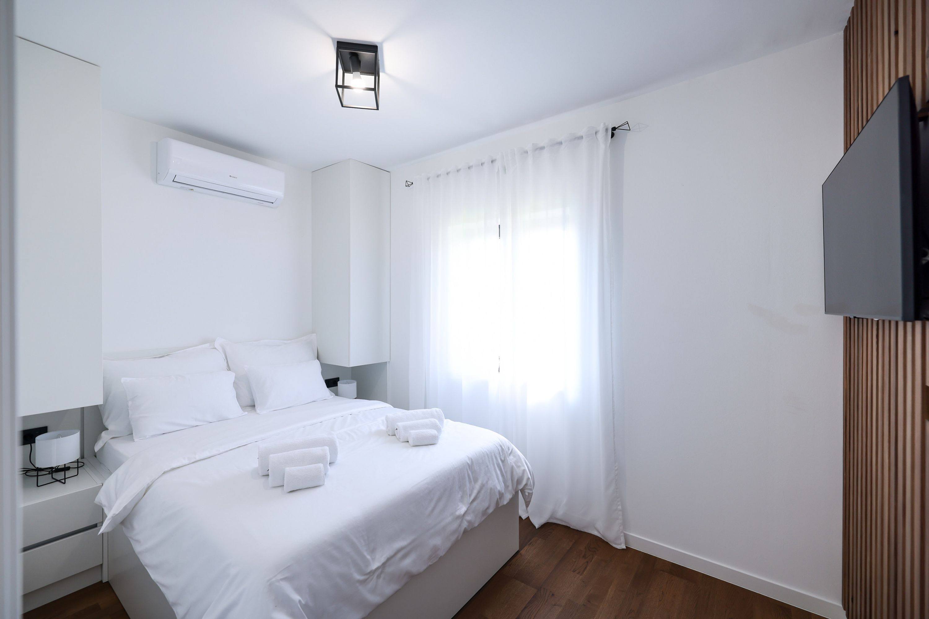 A hotel room with a bed, white curtains, a mounted TV, and a ceiling air conditioner.