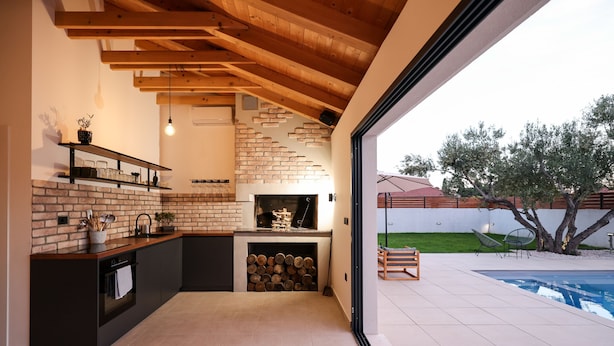 Eine moderne Küche mit freiliegenden Ziegelwänden, Holzbalken und einem großen Fenster mit Blick auf einen Swimmingpool.