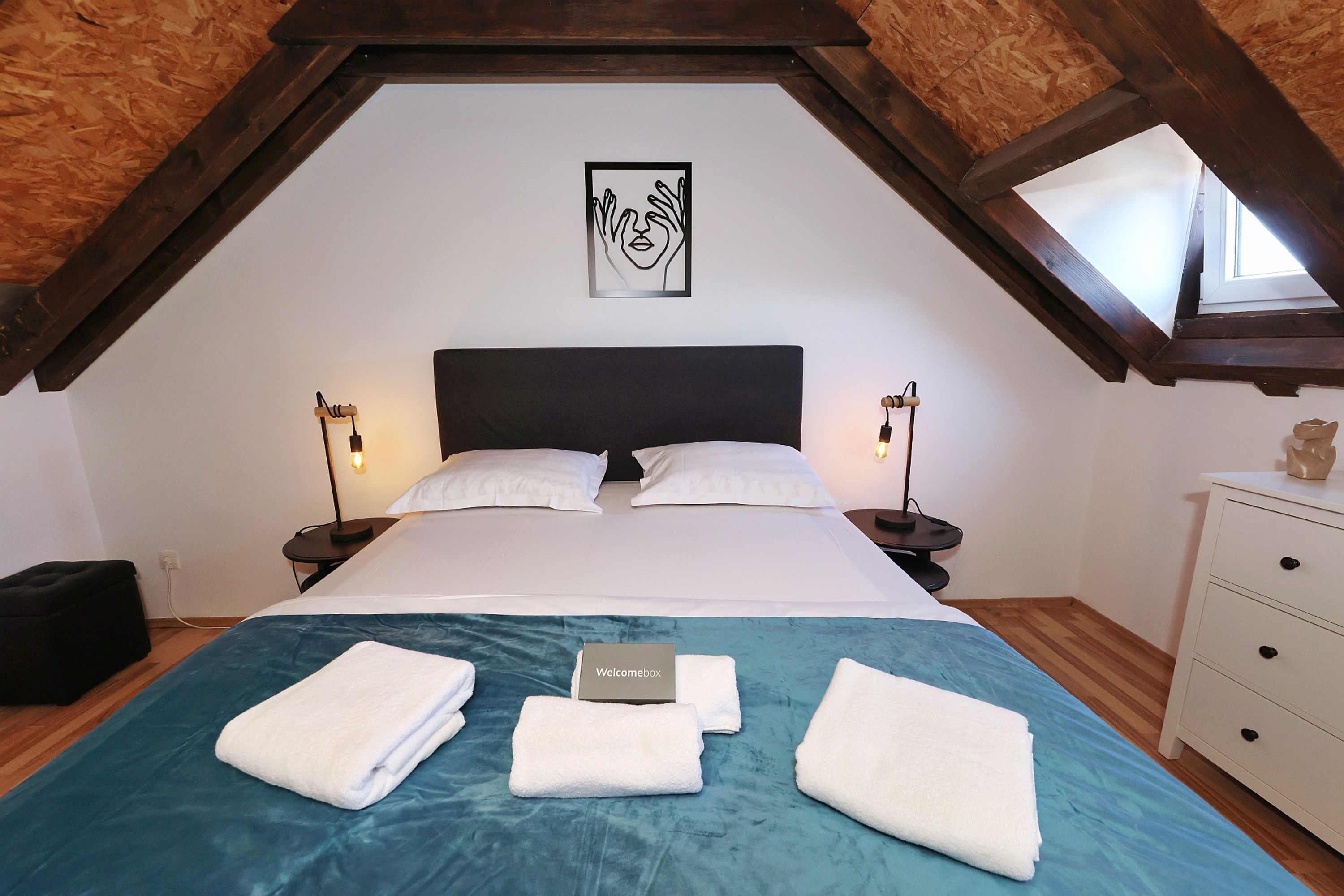 A bedroom with a sloped ceiling, a bed with white linens, a blue bedspread, and a welcome box on the bed.