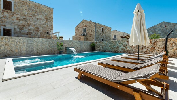 A swimming pool with lounge chairs and a waterfall feature, surrounded by stone buildings. - Villa Rea 3 With Heated Pool (Privlaka)
