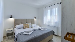 A modern bedroom with a bed, bedside tables, a nightstand, a window with sheer curtains, and a wooden bench. - Villa Rea 3 With Heated Pool (Privlaka)