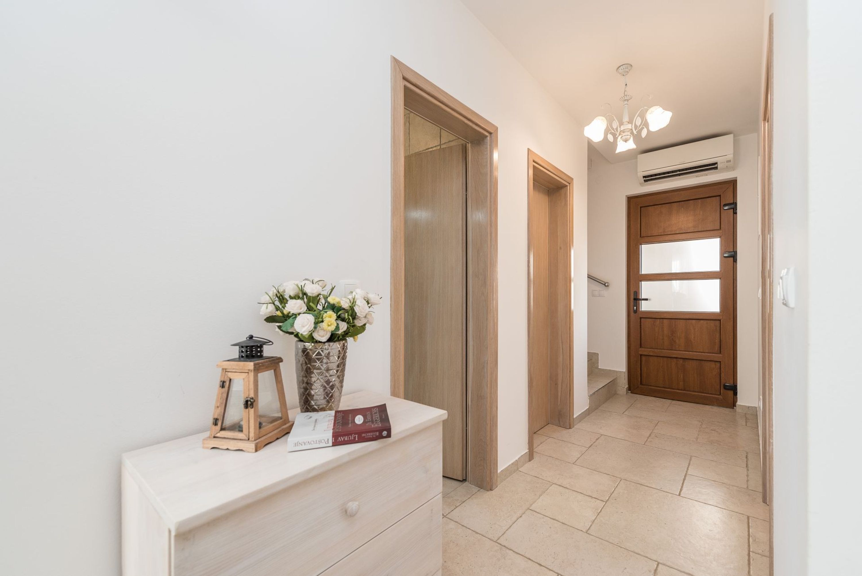 Un pasillo con puerta de madera, pared blanca, una pequeña mesa con un jarrón de flores y una lámpara en el techo.