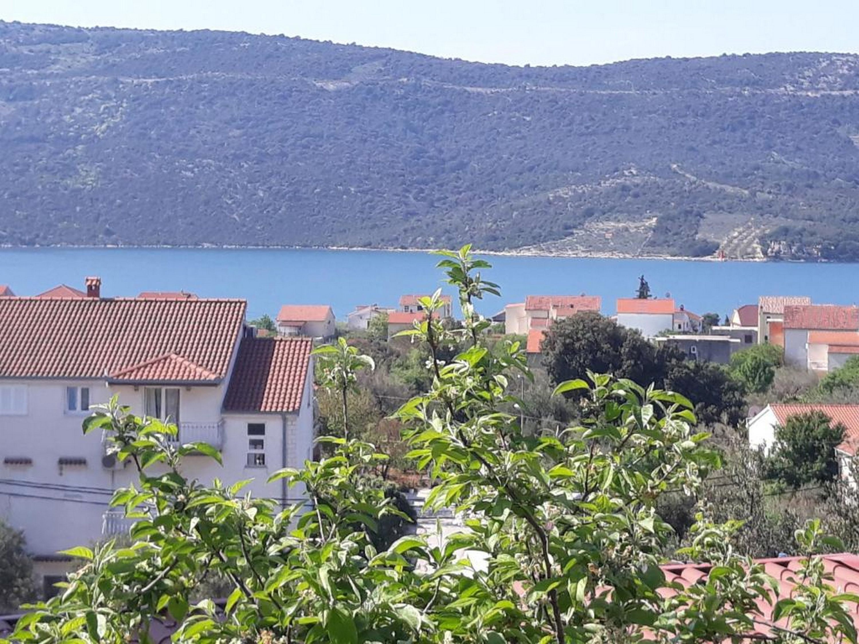 Un village côtier aux bâtiments aux toits de tuiles rouges, bordé par une mer d’un bleu limpide, avec des collines en arrière-plan.