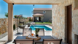 A poolside area with a wooden deck, a stone wall, and a view of a house with a tiled roof. - Villa Rea 4 With Heated Pool (Privlaka)