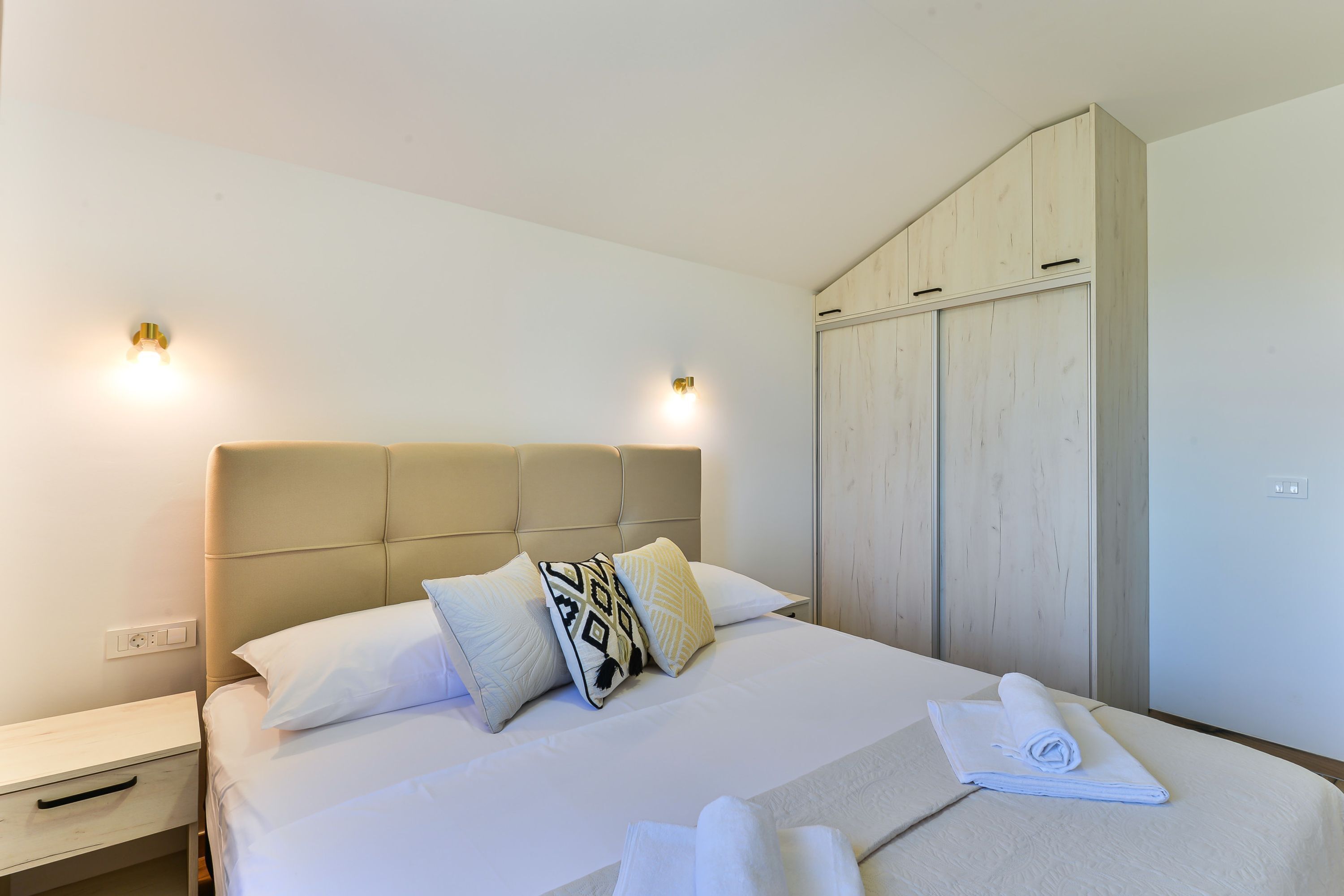 A neatly made bed with white linens and patterned pillows, a bedside table with a drawer, and a wooden wardrobe against the wall.