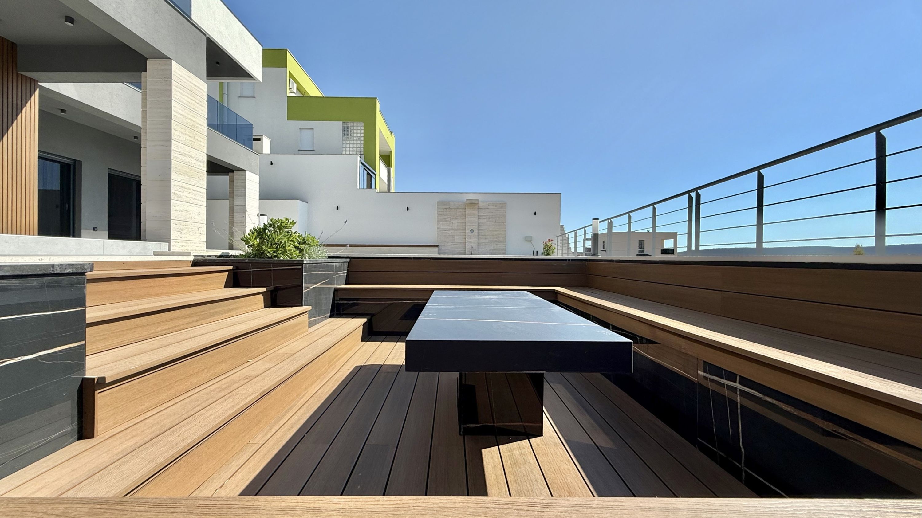 Terraço no telhado com bancos de madeira e uma mesa preta, com vista para um edifício moderno com detalhes verdes.
