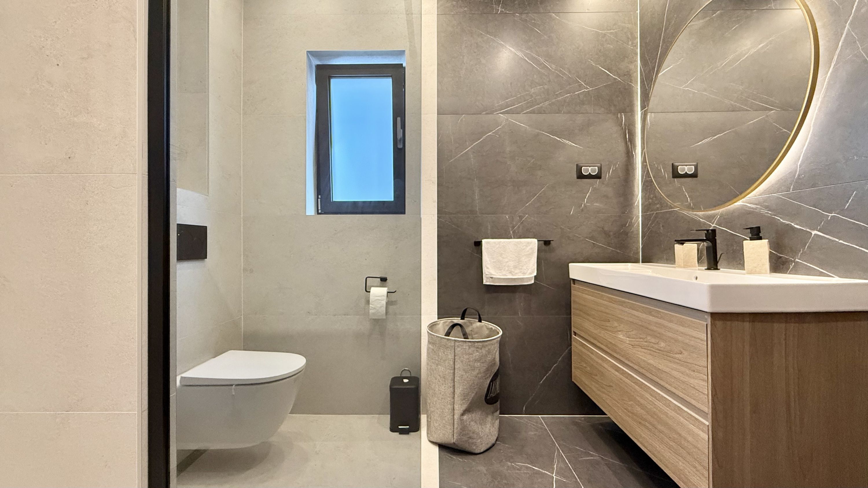 Een moderne badkamer met een toilet, een klein raam, een handdoekhouder, een wastafel met spiegel en een prullenbak.