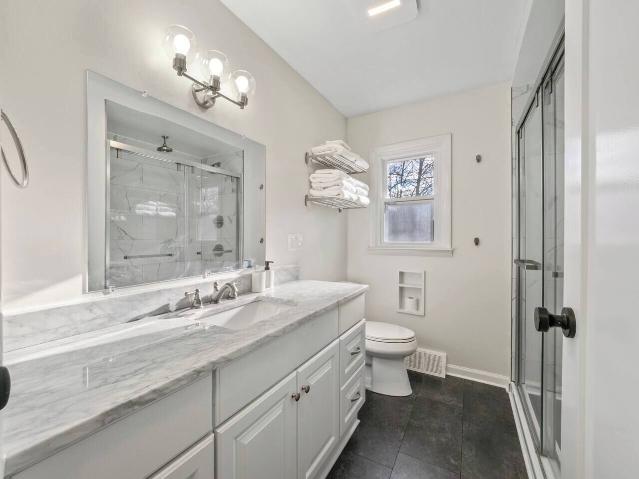 A modern bathroom with a large mirror, a sink with a marble countertop, a toilet, and a glass shower enclosure.