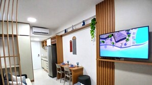 A modern hotel room with a bed, a TV mounted on the wall, a desk with two chairs, and a kitchenette in the background. - Mahrak Villa - Porto de Galinhas (Ipojuca)
