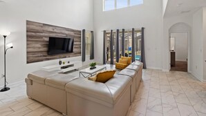 Een ruime woonkamer met een grote hoekbank, een flatscreen televisie bevestigd aan een houten wandpaneel en een groot raam dat zorgt voor natuurlijk licht.