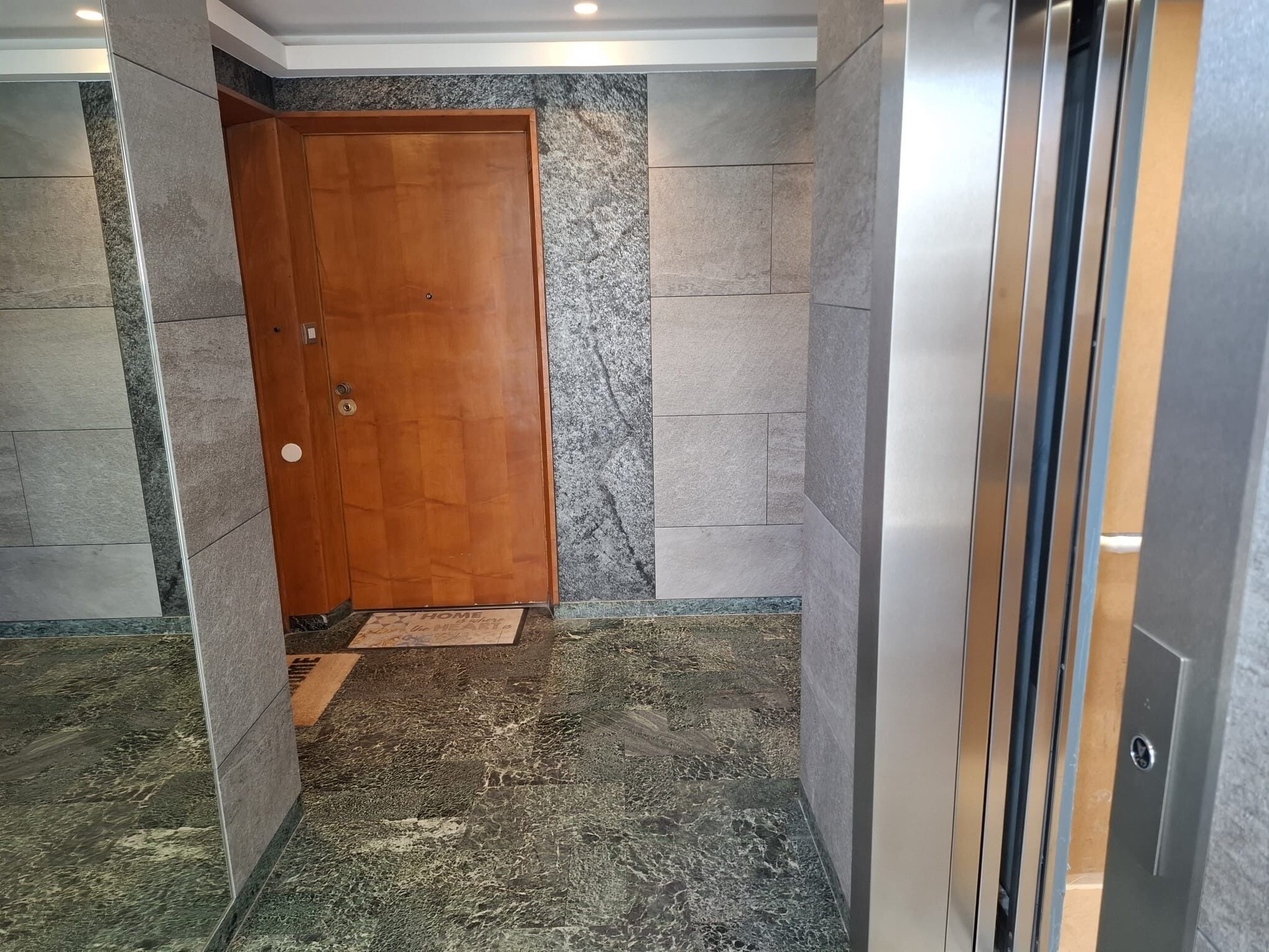 Um corredor com porta de madeira, paredes de mármore e um elevador metálico.