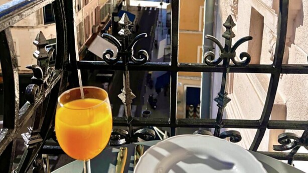 Un balcon avec un verre de jus d’orange, une assiette blanche et une vue sur une rue côtière.