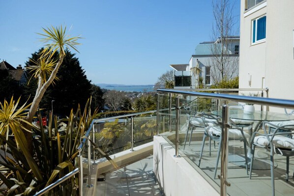 A balcony with glass railings overlooking a coastal view, featuring a dining area with chairs and a table.