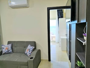 Ein Wohnzimmer mit grauem Sofa, zwei gemusterten Kissen, einer weißen Klimaanlage und Blick auf einen Balkon durch eine offene Tür.