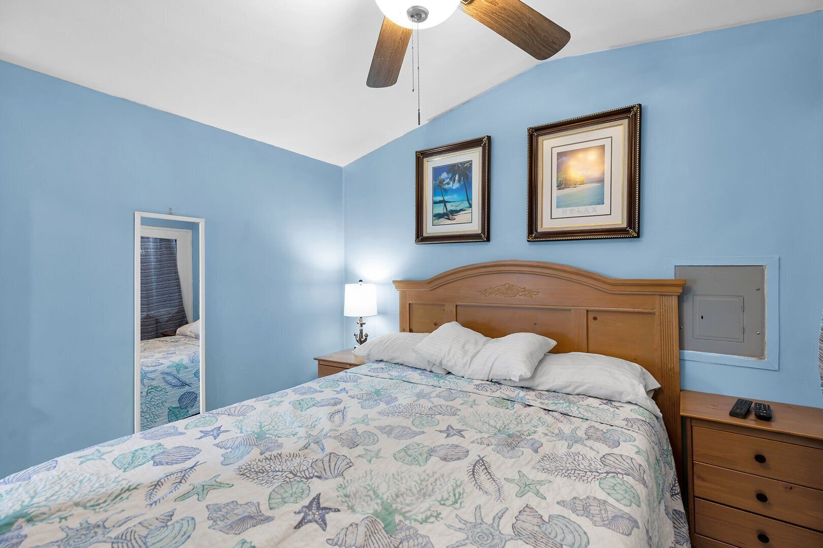 Quarto com cama, mesa de cabeceira, luminária, ventilador de teto e duas imagens emolduradas na parede.