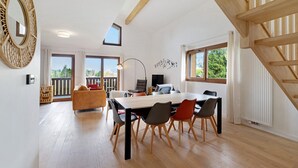 A spacious room with a wooden dining table, chairs, a sofa, and a large mirror. There is a balcony with a view of greenery.