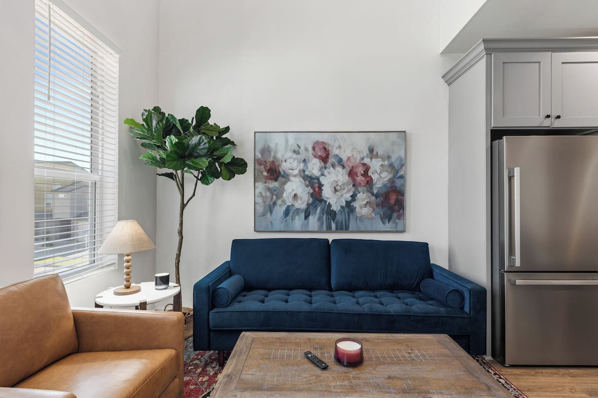 A living room with a blue sofa, a brown armchair, a coffee table with a remote control and candle, a large plant, and a painting on the wall.