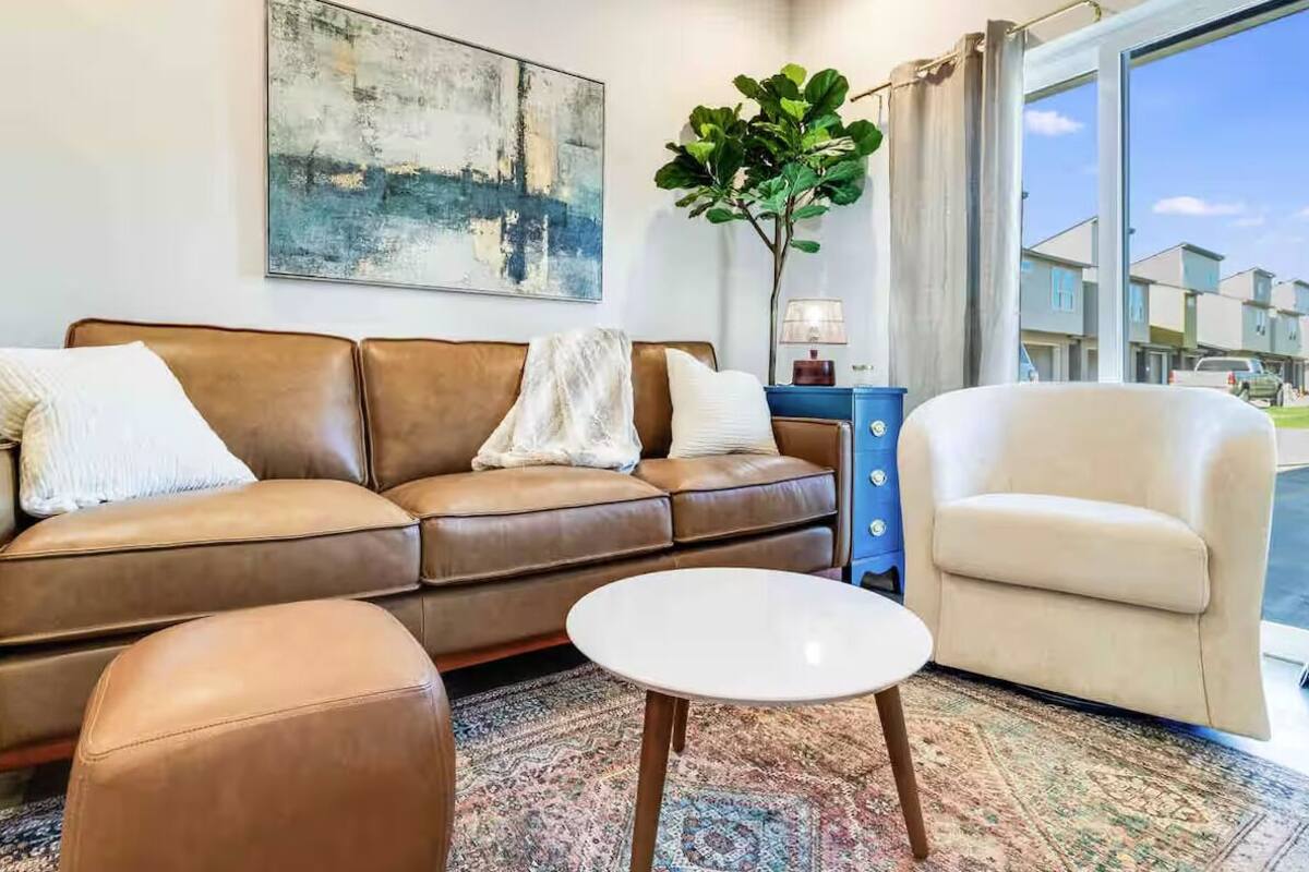A living room with a brown leather sofa, a white armchair, and a small white coffee table. There is a large abstract painting on the wall and a plant in the corner.