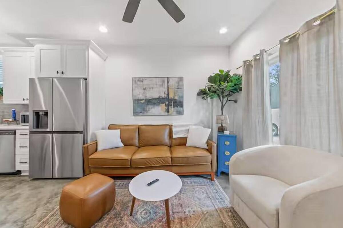 A modern living room with a brown leather sofa, a white coffee table, and a beige armchair. There is a kitchen area with stainless steel appliances and a blue cabinet. A painting hangs on the wall, and a plant is on a blue cabinet.