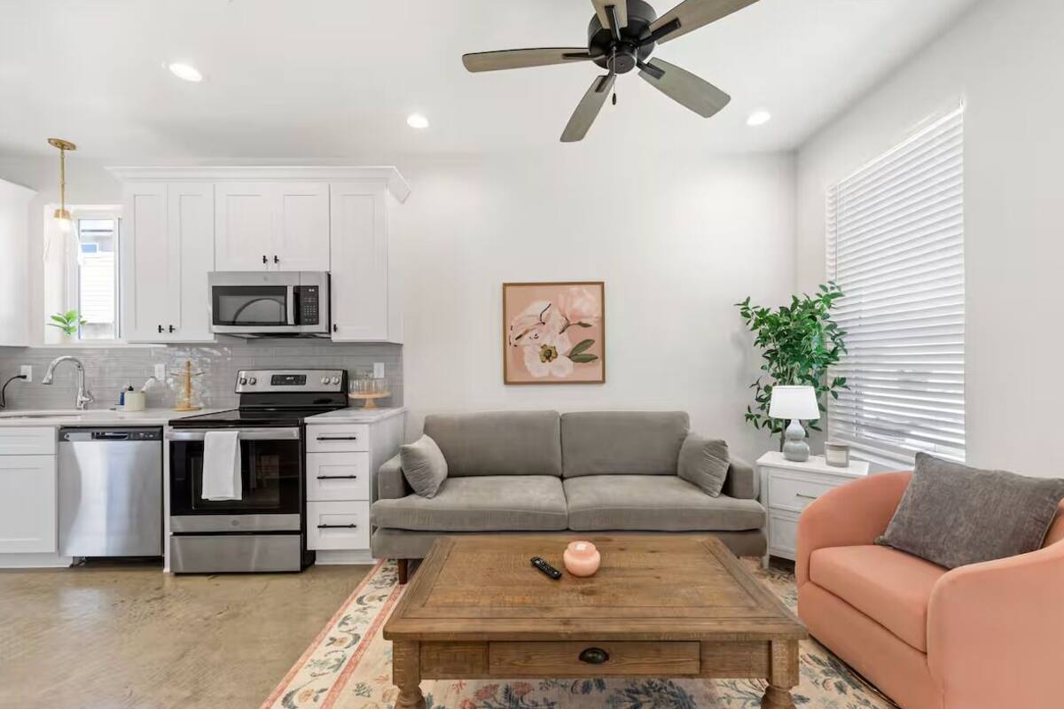 A modern kitchen with white cabinets, a microwave, and an oven. There is a gray sofa and a wooden coffee table in the living area.
