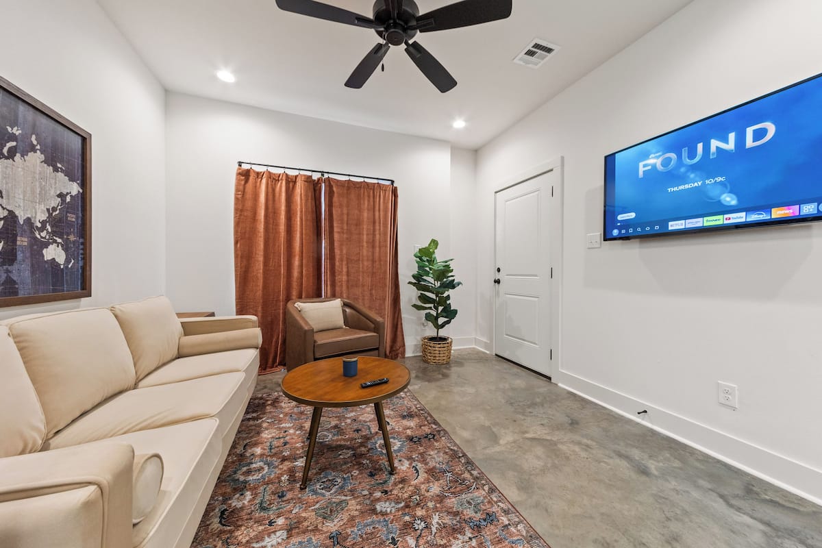 A modern living room with a beige sofa, a round coffee table, a large flat-screen TV mounted on the wall, a ceiling fan, and a large world map on the wall.