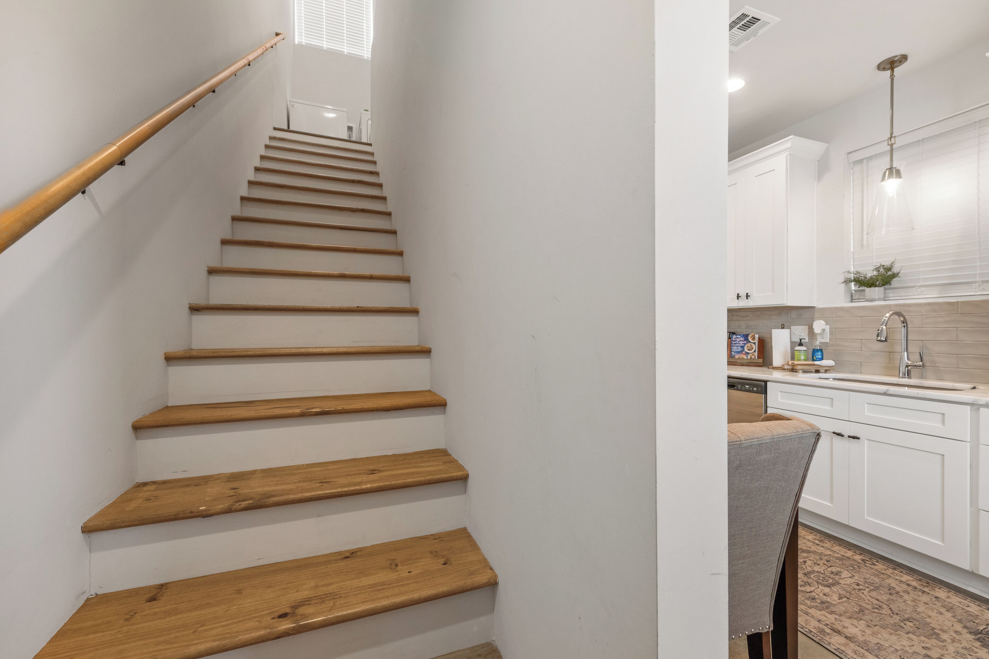 Un escalier en bois mène à une cuisine dotée de meubles blancs et d’une fenêtre.