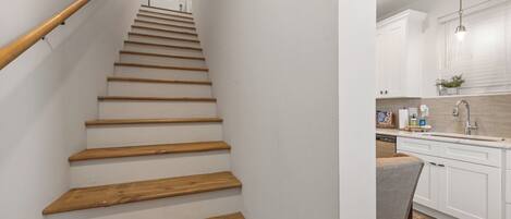 Un escalier en bois mène à une cuisine dotée de meubles blancs et d’une fenêtre.