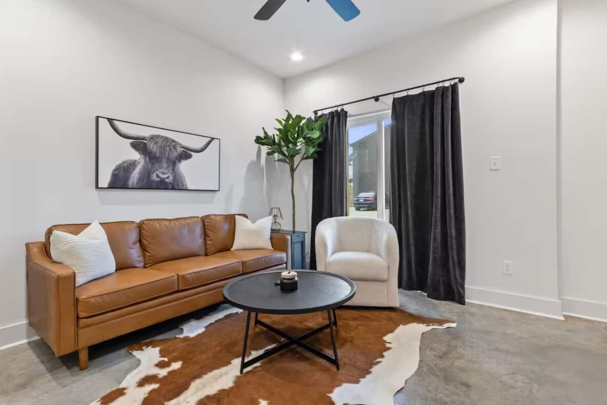A modern living room with a brown leather sofa, a round coffee table, a white armchair, and a large black and white cowhide rug.