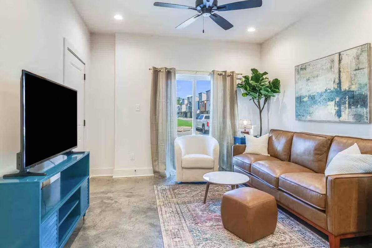 A modern living room with a flat-screen TV, a leather sofa, a coffee table, and a large window with curtains.