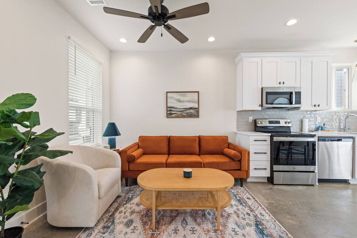 A modern kitchen with white cabinets, a microwave, stove, and dishwasher. There's a beige sofa, a wooden table with a blue mug, and a large plant.