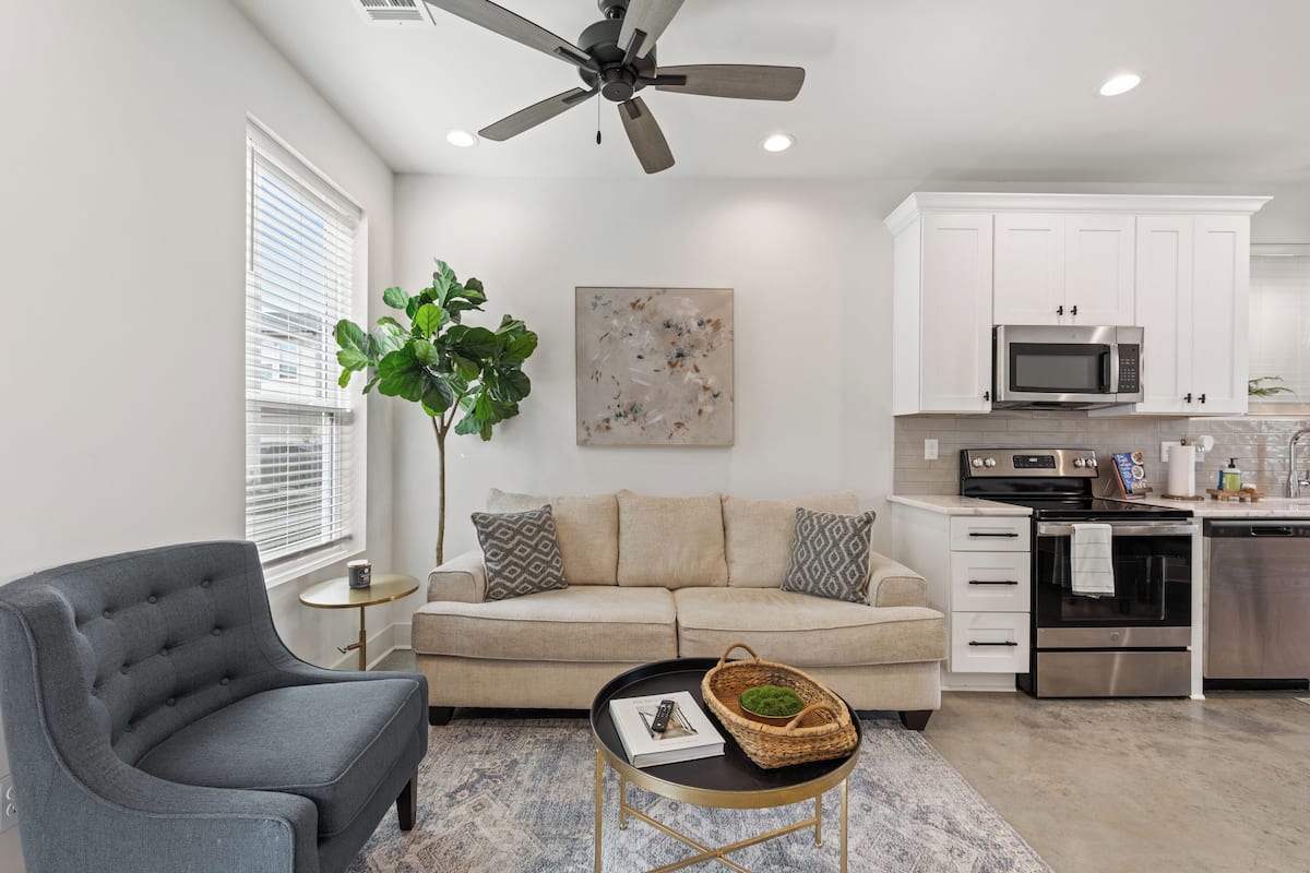 A modern living room with a grey armchair, beige sofa, and a small round table with a book and a basket. A large green plant is beside the sofa. The kitchen features white cabinets, a microwave, oven, and dishwasher.